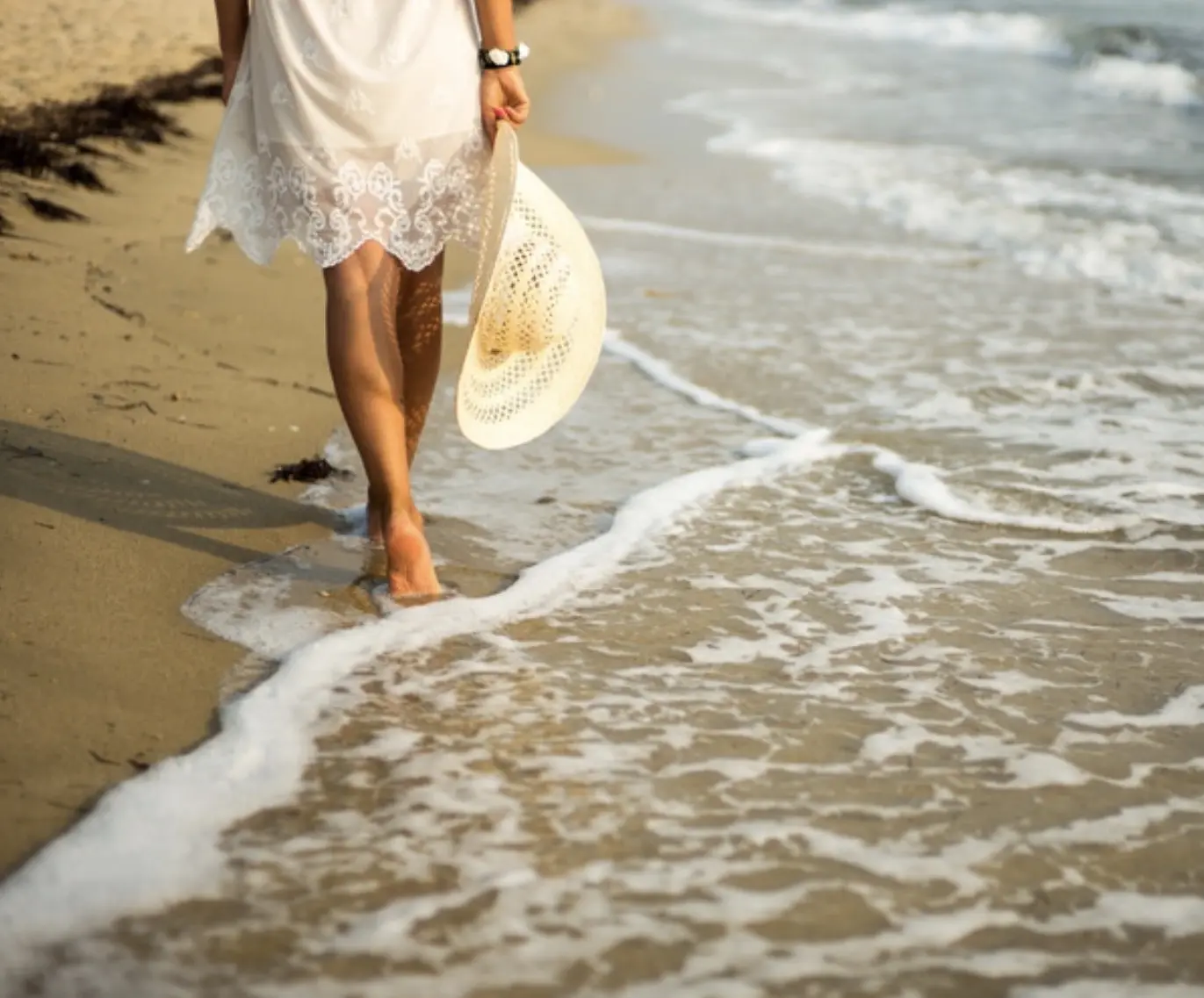 Una mujer camina descalza por la orilla, sosteniendo un sombrero de playa junto al mar